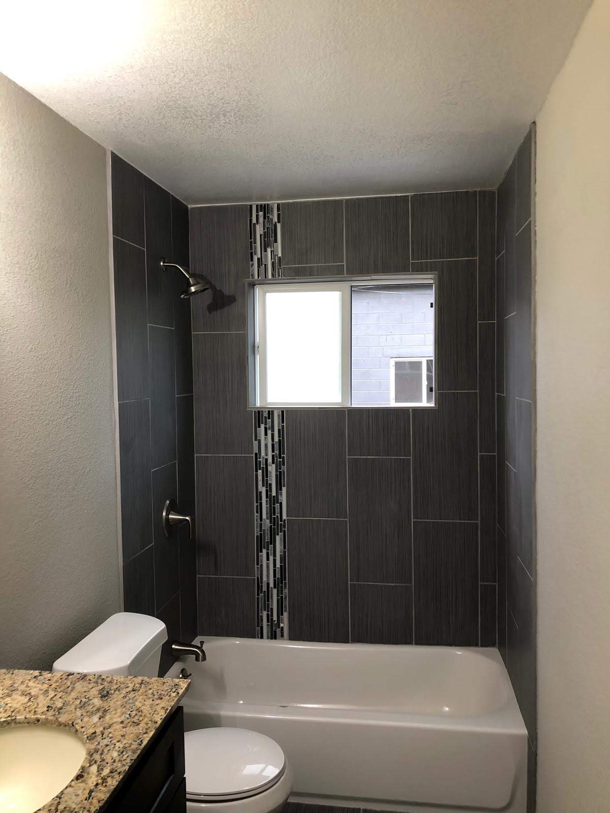 Bathroom remodel with a dark tiled tub surround, vertical mosaic accent, window, and granite vanity